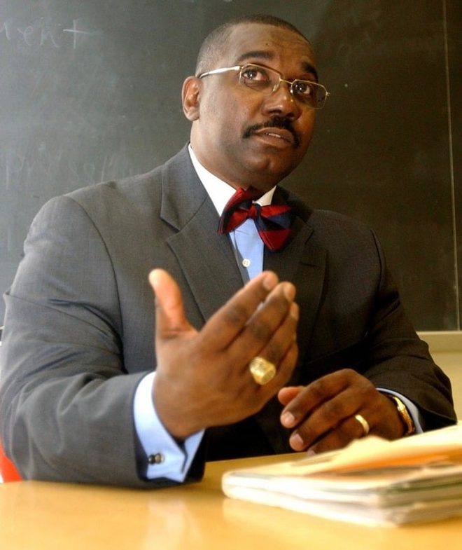 Raymond Edward Janifer, PhD, seated at a desk in front of a chalkboard, wearing a gray suit and red-and-navy bow tie